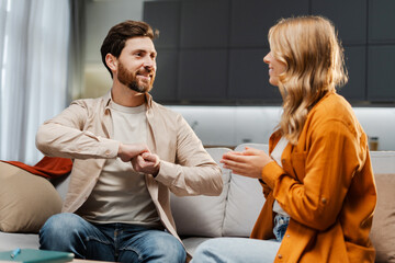 Fototapeta premium Portrait of positive happy young couple, smiling woman and man sitting on comfortable sofa at home, communicating with sign language, explaining. Communication concept