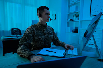 Military service. Soldier in headphones working on computer at wooden table in office at night