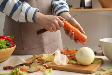 Woman peeling fresh carrot with knife at table in kitchen, closeup