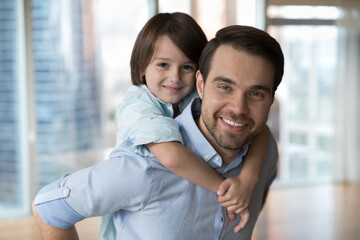 Close up portrait little pretty boy cuddles young father, feeling love enjoy time together. Man piggyback preschool son smile look at camera posing in modern apartment. Happy father Day, family ties