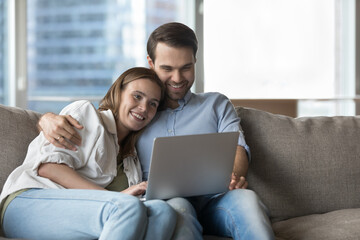 Fototapeta premium Couple in love resting on sofa watch online movie on laptop. Spouses spend carefree weekend on cozy couch, video calling to family using application, choose goods for home buying in e-commerce website