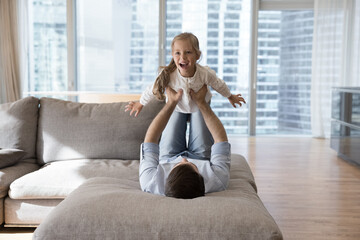 Father enjoy playtime with little daughter, man lying on sofa lifts kid girl on arms cute child...