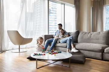 Foto op Plexiglas Carefree couple relaxing on sofa, little daughter play wooden cubes seated at table on floor, family spend pastime in modern apartment in skyscraper building. Leisure, new home, bank loan, tenancy © fizkes