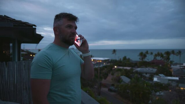 Bearded positive healthy guy standing on balcony with tropical island views on overcast day. Athletic smiling guy talking outside using smartphone. Businessman free lancer working remotely answer call