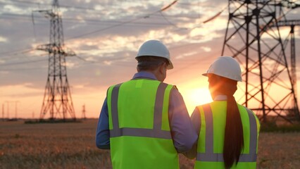 Electrician with tablet shows electric power transmission lines to inspector in sunset field...