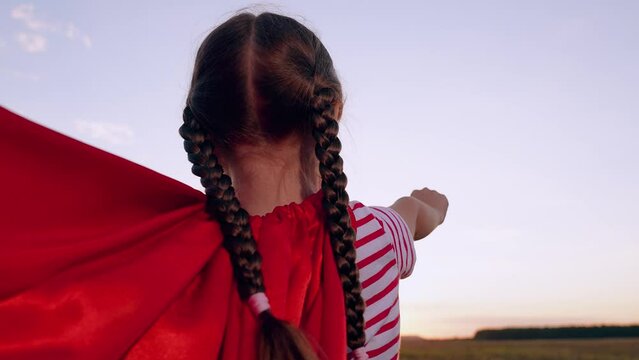 Cute little girl superhero flying in red cloak sunset sunrise countryside field closeup back view slowmo. Female kid child playing hero costume superman fantasy imagination character universe protect