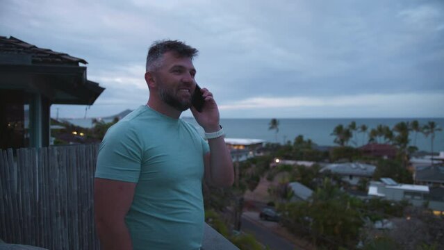 Athletic smiling guy talking outside using smartphone. Bearded businessman free lancer working remotely answer call. Positive healthy guy standing on balcony with tropical island views on overcast day