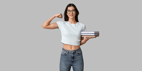 Young woman with books flexing muscles on grey background. Feminism concept