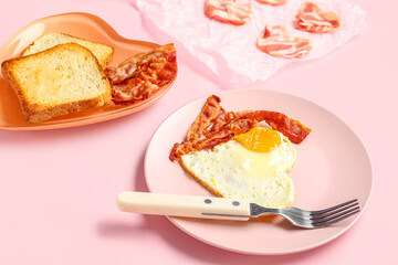 Plates with tasty bacon, heart made of fried egg and toasts on pink background, closeup