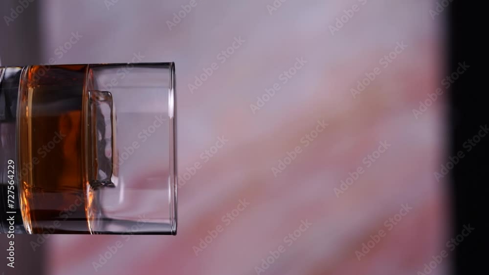 Poster glass of whiskey with an ice cube rotating on a circular platform vertical video