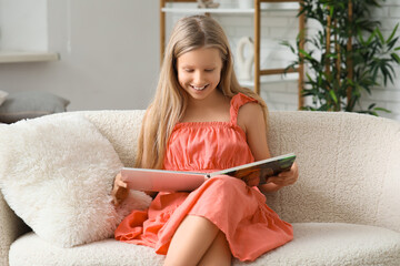 Cute little girl reading book while sitting on sofa at home