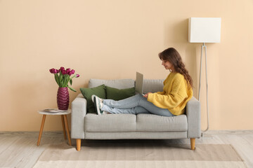 Pretty young woman using modern laptop and vase with bouquet of beautiful tulips at home