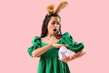 Surprised young woman in bunny ears with Easter egg on pink background