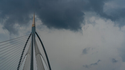 A fragment of the architecture of a cable-stayed bridge against a cloudy sky. A tall pole with a...