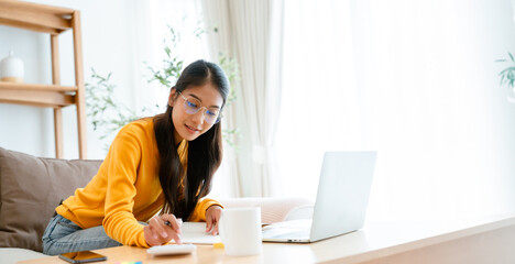 Young Asian female wearing glasses using laptop, working at home in living room, coffee mug on...