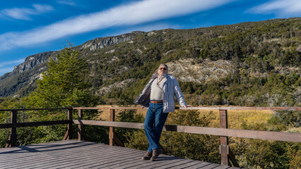 Fototapeta na wymiar A man stands on a plank observation deck, leaning on the railing. Behind him is the beautiful landscape of Patagonia. A mountain against a blue sky and clouds, green trees, grass in a meadow.Argentina