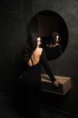 Sexy woman in a black dress posing near a mirror in a dark room. Fashion shooting concept