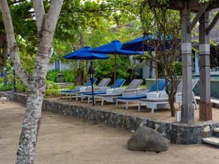 Rows of poolside and outdoor lounge chairs with blue and white color theme and placed strategically surrounding the pool and by the beach sand near a beach bar.