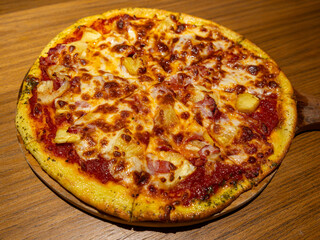 A mouth watering hawaiian pizza with common toppings plus slices of pineapples served on a wooden brown table under a warm spot light.