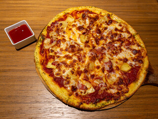 A mouth watering hawaiian pizza with common toppings plus slices of pineapples served on a wooden brown table under a warm spot light.