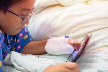 Obraz na płótnie Canvas Little Asian boy illness lay on sickbed in hospital with a saline intravenous (IV) on hand holding smartphone and watching on youtube.