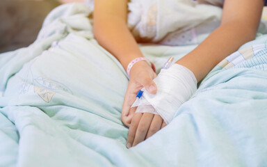 Young patient asian boy sitting on bed in hospital with IV saline drip to back of the hand,...