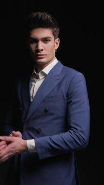 mysterious young businessman in blue suit walking, being confident, rubbing palms and looking to side in front of dark background