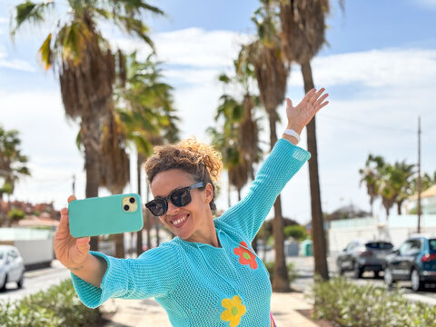 One excited woman taking selfie outdoor with blue mobile phone. Street with palms and car in background. One female tourist enjoy leisure activity alone and online connection. People happiness day