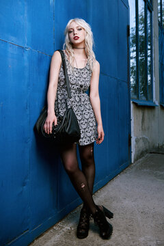 Pretty young grunge (rock) girl in dress, ripped pantyhose and sunglasses with bag in hand standing at wall. Full length outdoor portrait of informal model