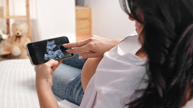 Happy pregnant woman lying on sofa and viewing child sonogram on display of smartphone, baby picture