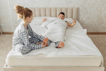 the children are wrapped in a blanket and tickling each other's heels on the bed. Family having fun at home. Leisure - obrazy, fototapety, plakaty