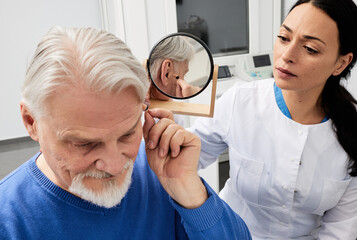 Senior man trying on hearing aid in front of mirror with female audiologist, fitting hearing device...