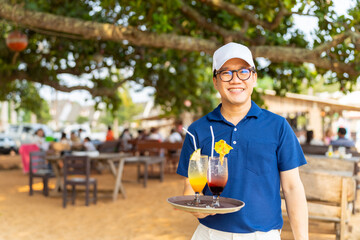 Portrait of Asian man waiter serving food and drink to customer on the table at tropical beach cafe and restaurant on summer holiday vacation. Food and drink business service occupation concept. - obrazy, fototapety, plakaty