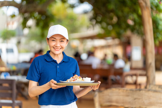 Portrait of Asian woman waitress serving food and drink to customer on the table at tropical beach cafe and restaurant on summer holiday vacation. Food and drink business service occupation concept.