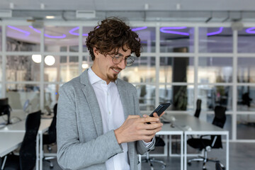 A young man works in an office, stands inside and uses a mobile phone with a smile, typing messages, texting.