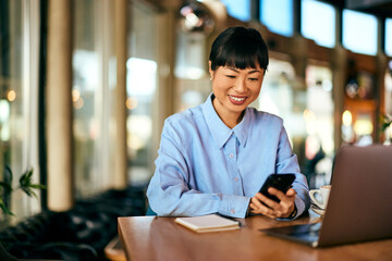A freelance Asian woman sitting at the cafe, using a mobile phone, working online.