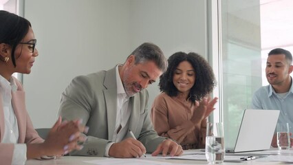 Happy old businessman executive investor banker signing legal business document agreement contract at board meeting with attorneys lawyers advisers diverse law team applauding sitting at office table.