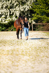 Young woman with a black shirt and short highlighted hair stands with her horse on a sunny riding arena.