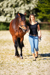 Young woman with a black shirt and short highlighted hair stands with her horse on a sunny riding arena.