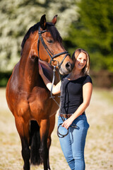 Young woman with a black shirt and short highlighted hair stands with her horse on a sunny riding arena.