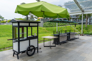 A wooden black and white push cart placed along side with two minimalist food stalls under a green...