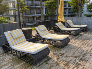 Four outdoor chaise on a wooden pool deck floor in front of a residential building on a sunny...