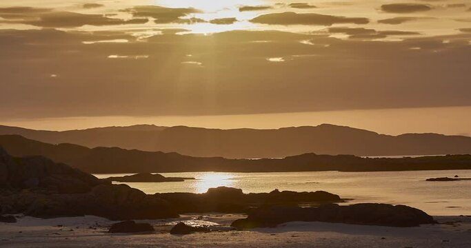 Time lapse sequence of setting sun behind clouds at the rocky shoreline of Fidden Beach, Isle of Mull, Inner Hebrides, Scotland, United Kingdom