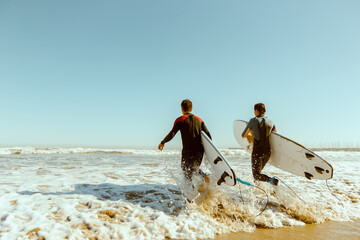 Back view of group of friends with surfboards in wetsuit entering towards ocean for surfing on waves - obrazy, fototapety, plakaty