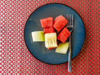 A few chunks of red watermelon and honeydew melon cubes served in a blue ceramic plate.