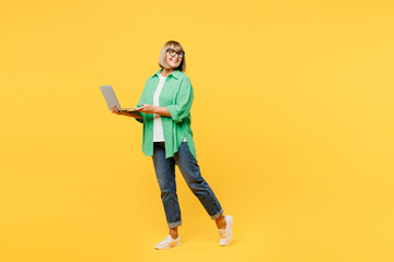 Full body elderly blonde woman 50s years old wear green shirt glasses casual clothes hold use work on laptop pc computer look aside walk isolated on plain yellow background studio. Lifestyle concept.