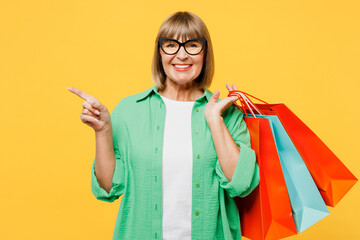 Elderly woman 50s years old wears green shirt casual clothes hold in hand paper package bags after shopping point finger aside isolated on plain yellow background. Black Friday sale buy day concept.