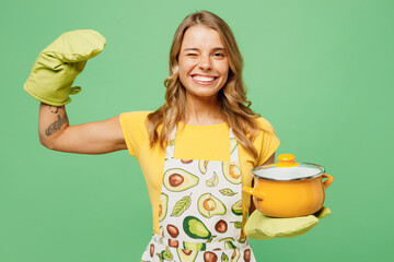 Young fun housewife housekeeper chef cook baker woman wear apron yellow t-shirt hold in hand pot pan show muscles wink blink eye isolated on plain pastel green background studio. Cooking food concept.