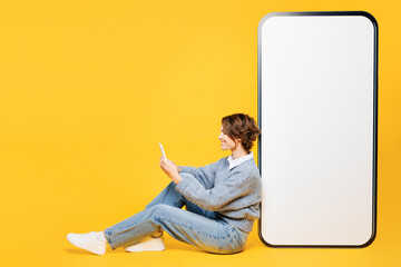Full body young woman wears grey knitted sweater shirt casual clothes sit near big huge blank screen mobile cell phone with area use smartphone isolated on plain yellow background. Lifestyle concept. - obrazy, fototapety, plakaty