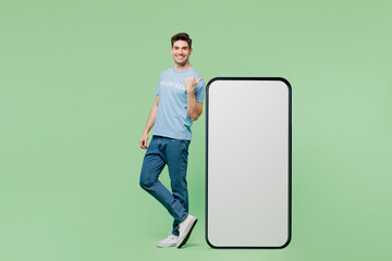Full body young man wear blue t-shirt title volunteer point finger on big huge blank screen area mobile cell phone isolated on plain green background. Voluntary free work help charity grace concept.